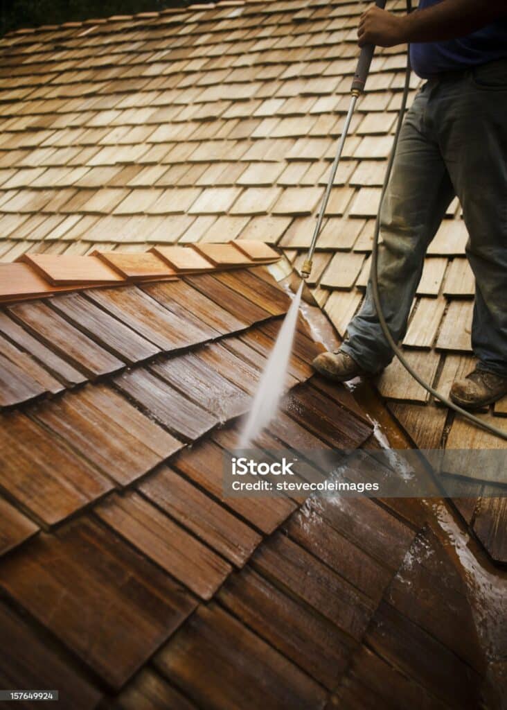 Man stands atop a roof while spraying the wooden shingles clean. Vertical shot.